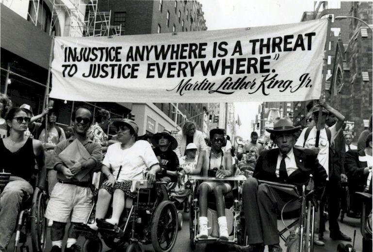 Group of disabled protesters at the NYC Disability Pride March holding a banner that reads "Injustice Anywhere is a Threat to Justice Everywhere." Dr. Martin Luther King.