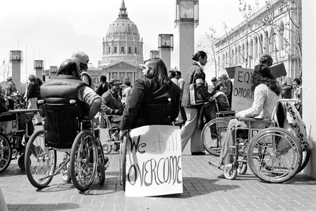 Black and white photograph taking in April 1977 by Anthony Tusler showing disabled protesters participating in the "504 Sit-In" in front of the SF Federal Building with signs attached to their wheelchairs that read "We Shall Overcome."